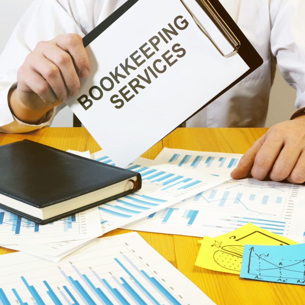 A person holding a clipboard labeled 'Bookkeeping Services' surrounded by charts and graphs on a desk, representing bookkeeping virtual assistant services.