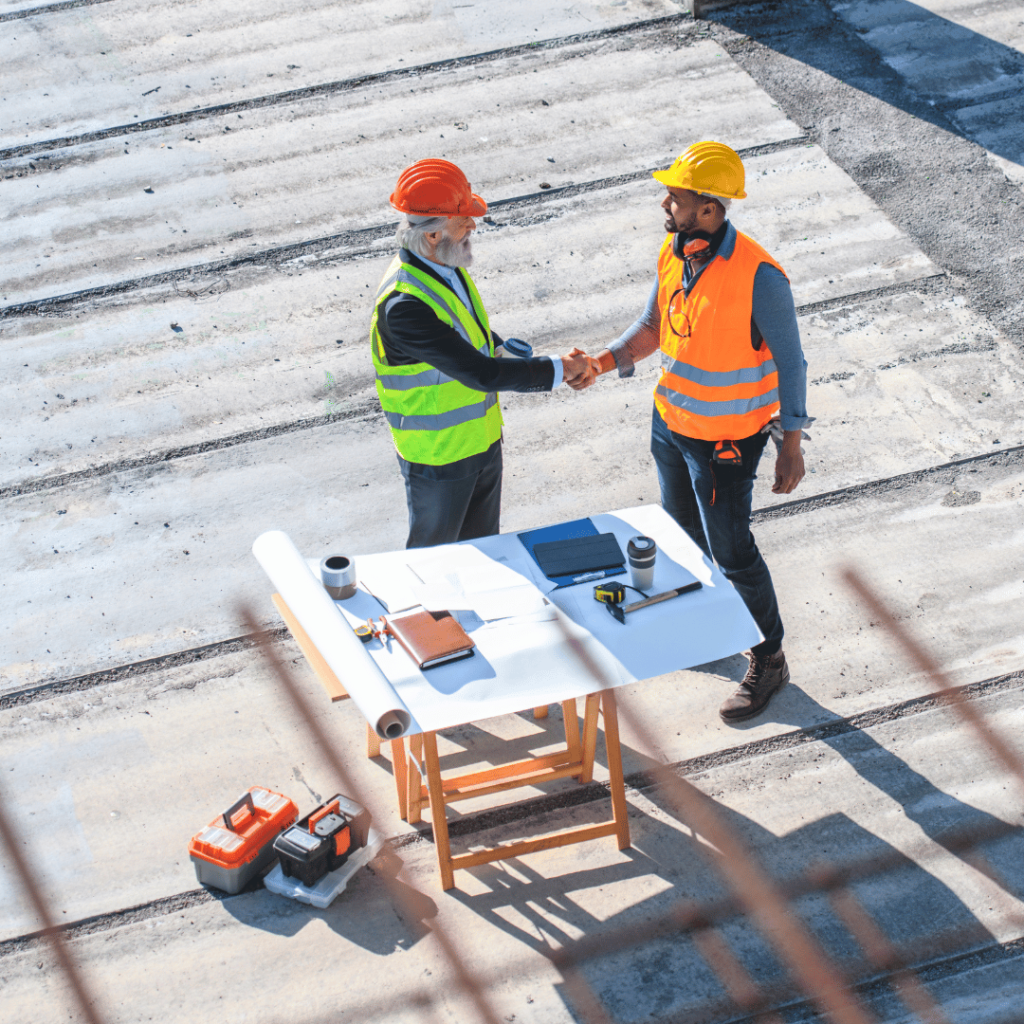 Two construction workers in safety gear shaking hands over a table with blueprints and tools on a construction site, representing performance-focused onsite services.