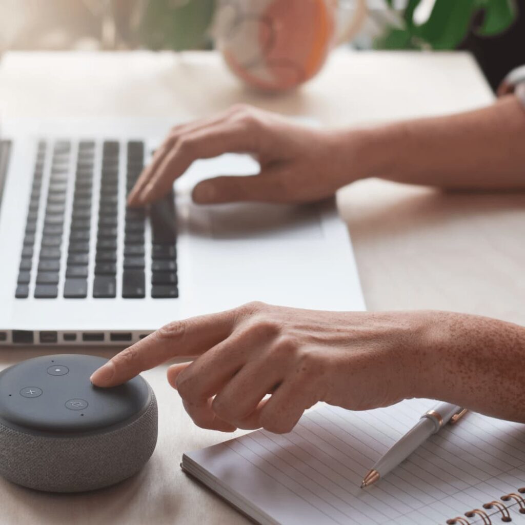 A person using a laptop and interacting with a smart speaker while taking notes, representing virtual personal assistant services